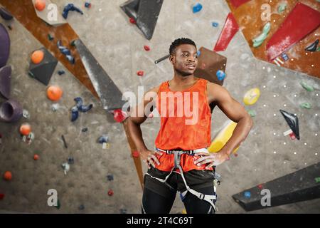 atletico afro-americano in camicia arancione con imbracatura alpina in posa con le mani sui fianchi Foto Stock