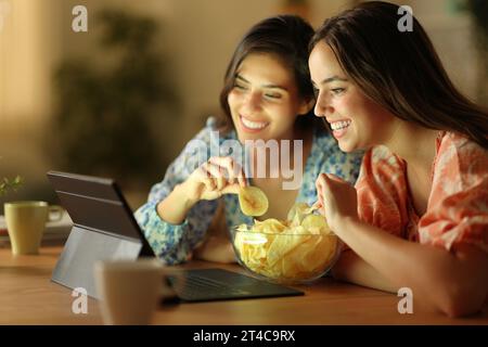 Due amici felici che guardano un film su un tablet mentre mangiano patatine fritte la sera a casa Foto Stock