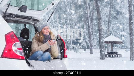 Donna con cappello di lana siede nel bagagliaio dell'auto e tiene una tazza di tè caldo in mano. Foto Stock