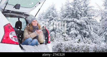 Donna con cappello di lana siede nel bagagliaio dell'auto e tiene una tazza di tè caldo in mano. Foto Stock