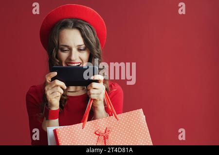 Buon Valentine. felice donna moderna di 40 anni in abito rosso e berretto isolato su sfondo rosso con borse della spesa utilizzando applicazioni per smartphone e. Foto Stock