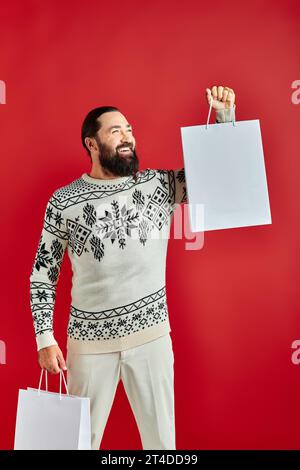 Uomo con barba positiva in maglione natalizio che regge borse dello shopping su sfondo rosso, vendite natalizie Foto Stock