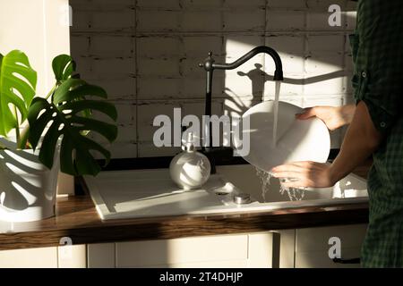 le mani di una donna lavano i piatti a casa. Piatti sotto un flusso d'acqua alla luce del sole. routine domestica. Foto Stock