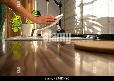le mani di una donna lavano i piatti a casa. Piatti sotto un flusso d'acqua alla luce del sole. routine domestica. Foto Stock