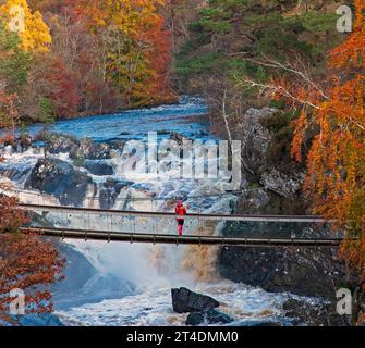 Rogie Falls, Strathpeffer Highland, Scozia, Regno Unito. 30 ottobre 2023. il sole nuvoloso sopra la scena autunnale prima di una forte pioggia. Nella foto: Una donna vestita con colori vivaci che si trova sul ponte sospeso, che contrasta con lo sfondo, scatta una foto al telefono cellulare della splendida vista sul fiume BlackwaterRiver. Foto Stock