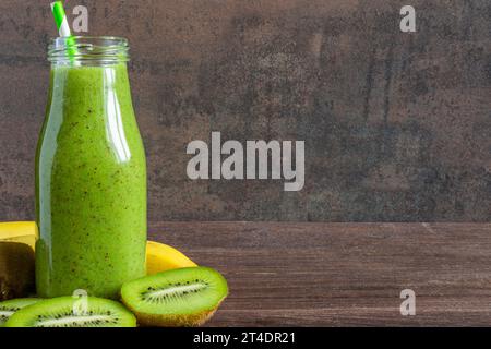 Frullato verde a base di kiwi e banana in bottiglia di vetro con frutta fresca su fondo rustico di legno. Dieta sana bevanda Foto Stock