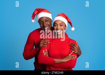 Felici sposi neri con cappelli Babbo Natale che si abbracciano su sfondo blu Foto Stock