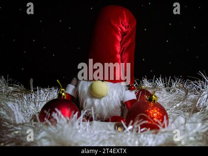 Uno gnomo divertente di Capodanno con un cappello rosso circondato da decorazioni rosse dell'albero di Natale e tinsel bianco sullo sfondo dell'imitazione della sta Foto Stock