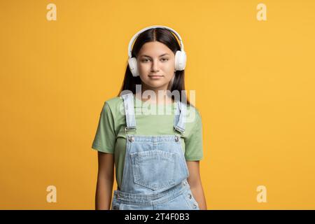 Ragazza amante della musica con le cuffie che ascolta la musica guardando la fotocamera su sfondo giallo. Foto Stock