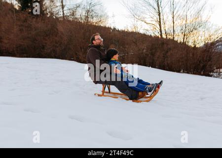 padre e figlia sorridenti insieme su una slitta di legno che scende sulla collina innevata Foto Stock