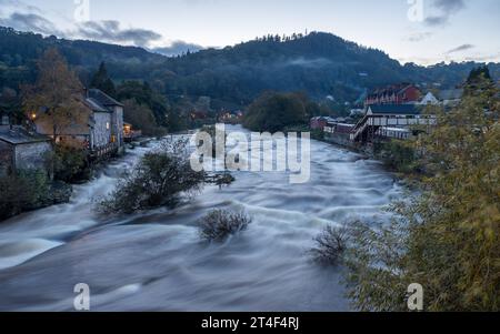 Un'immagine HDR suggestiva che guarda il fiume Dee a Llangollen al crepuscolo mentre le luci iniziano a brillare mentre la luce svanisce, vista il 22 ottobre 2023. Foto Stock