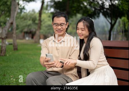 Una figlia asiatica felice e adorabile ama guardare video carini su uno smartphone con suo padre mentre si rilassa su una panchina in un parco insieme. Foto Stock
