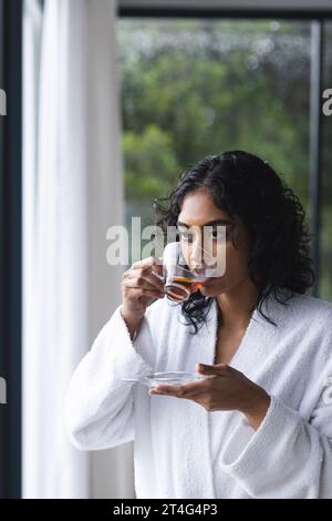 Felice donna birazziale in accappatoio che beve tè in una stanza soleggiata a casa Foto Stock