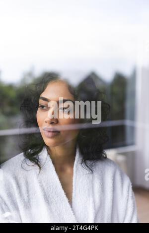 Donna birazziale pensiva in accappatoio che guarda fuori dalla finestra nella stanza soleggiata di casa Foto Stock