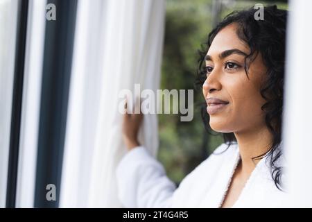 Donna birazziale pensiva in accappatoio che guarda fuori dalla finestra nella stanza soleggiata di casa Foto Stock