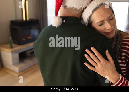 Felice coppia caucasica anziana che indossa i cappelli di babbo natale che si abbracciano a casa a natale Foto Stock