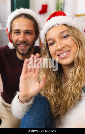 Felice coppia caucasica nei cappelli di natale con videochiamata, sorridendo e salutando a casa Foto Stock