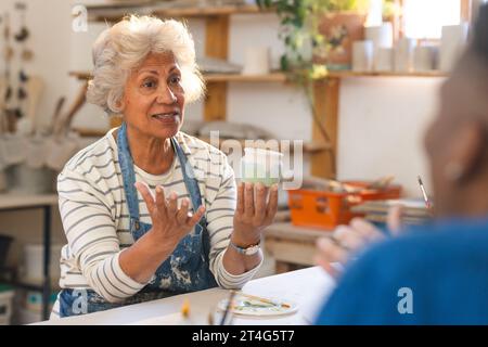 Felice ceramista birazziale anziana con i capelli grigi, discutendo con gli altri in studio di ceramica Foto Stock