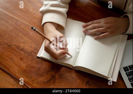 Immagine dall'alto di una donna che tiene in mano una matita, che scrive in un libro su un tavolo di legno. Mockup di pagine vuote vuote, mantenimento di diari, esercitazioni, creazione di elenchi Foto Stock