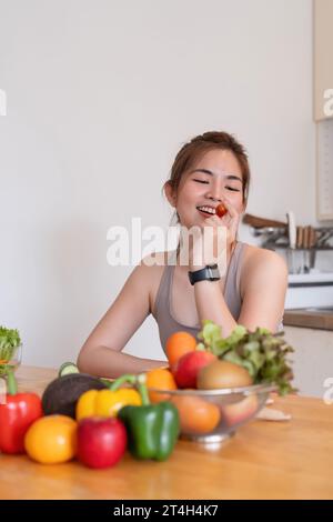 Giovane asiatico sana bella donna con abiti casual è sorriso e affettare pomodori tagliati per mangiare frutta a dieta a casa in cucina in vacanza Foto Stock