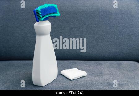 Prodotto per la pulizia con spugna. Smacchiatore per tessuti per mobili. Bottiglia bianca in plastica con detergente e tovagliolo su un divano grigio. Foto Stock
