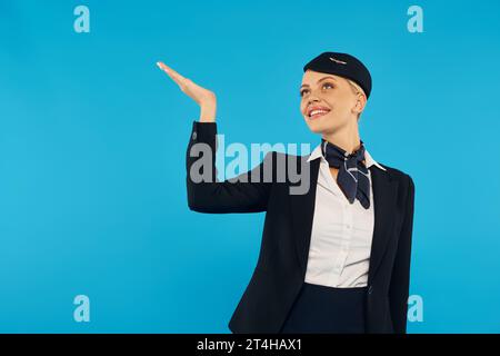 hostess eccitata ed elegante in uniforme aziendale che guarda verso l'alto e punta con la mano sul blu Foto Stock