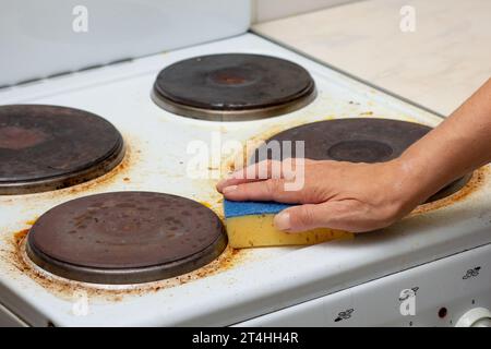 La mano di donna rimane macchiata di bruciature sulla sporca stufa elettrica bianca strofinando con una spugna Foto Stock