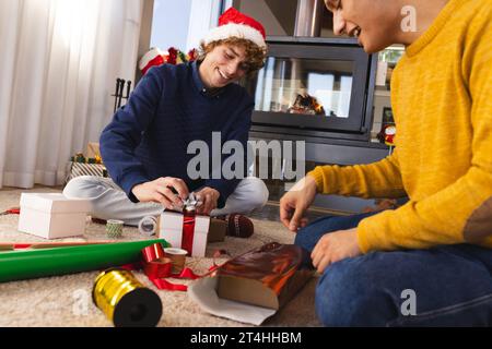 Felice coppia gay maschile eterogenea che indossa cappelli di natale e regali di imballaggio a casa Foto Stock