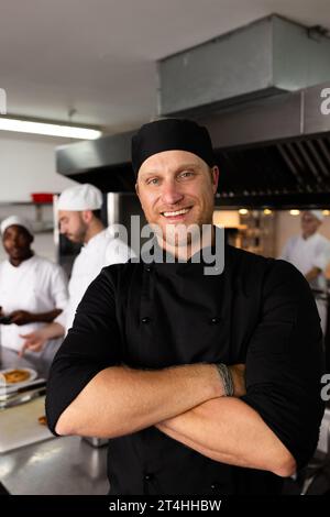 Chef maschio eterogeneo felice con le braccia incrociate in piedi contro gli studenti che cucinano in background Foto Stock