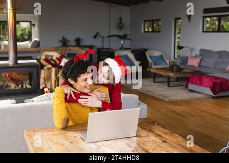 Felice coppia gay maschile eterogenea che si abbraccia indossando cappelli di natale e facendo videochiamata a casa Foto Stock