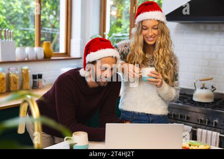 Felice coppia caucasica nei cappelli di natale che bevono un caffè e usano un computer portatile in cucina Foto Stock