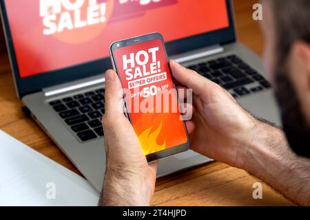 Uomo sul posto di lavoro che guarda annunci di vendita caldi sullo smartphone e sullo schermo del notebook. Fino al 50% di sconto. Foto Stock