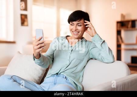 Sorridente donna di mezza età che naviga sui social media sul cellulare al chiuso Foto Stock