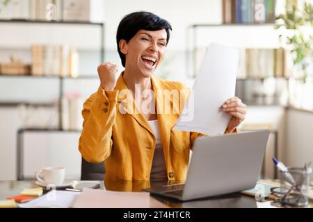 Donna d'affari europea che stringe pugni al computer portatile che tiene i documenti in ufficio Foto Stock