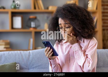 Donna triste delusa ha ricevuto notizie al telefono, donna afroamericana che legge cattive notizie online utilizzando l'app sullo smartphone, seduto a casa sul divano nel salotto di casa. Foto Stock