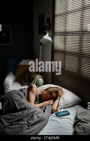 Una ragazza adolescente sconvolta sdraiata sul letto da una finestra coperta di veneziane la mattina presto e piange mentre soffre di cyberbullismo Foto Stock