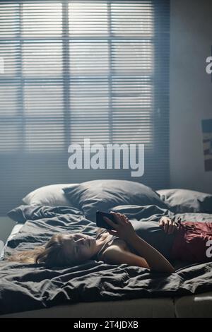 Una ragazza stressata che soffre di cyberbullismo mentre era sdraiata sul letto contro la finestra coperta da tende veneziane e guardava lo schermo dello smartphone Foto Stock