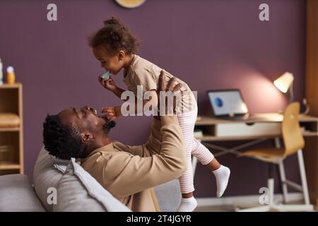 Ritratto laterale di un uomo afro-americano felice che gioca con la figlia piccola a casa e la tiene in aria sorridendo gioiosamente, copia spazio Foto Stock