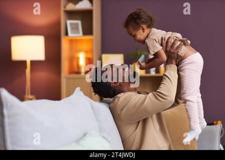 Vista laterale ritratto del giocoso padre Nero che si coccola con la figlia piccola a casa e la tiene in aria sorridendo gioiosamente, spazio copia Foto Stock