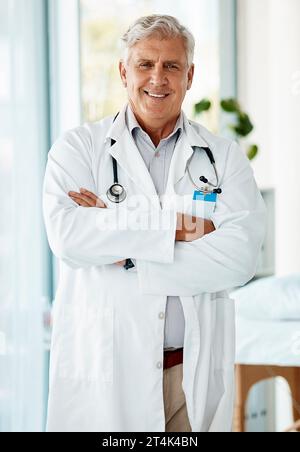 Esperto medico maschio maturo sorridente e in piedi con le braccia incrociate in un ospedale. Un uomo anziano ottimista che indossa un camice da laboratorio Foto Stock