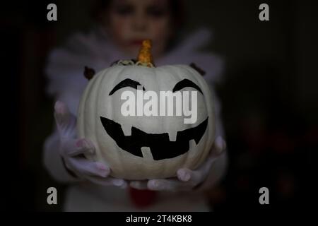 Zucca bianca sorridente di Halloween nelle mani di una ragazza vestita da spaventoso clown con illuminazione misteriosa Foto Stock