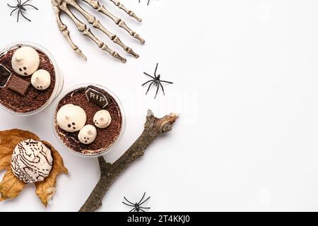 Tazze di plastica con deliziosi tiramisù, biscotti, mano scheletrica e ragni per festeggiare Halloween su sfondo bianco Foto Stock