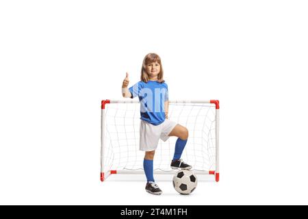 Ragazza in maglia sportiva in piedi con il piede sopra una partita di calcio e i pollici gestanti davanti a una rete di porta isolata su sfondo bianco Foto Stock