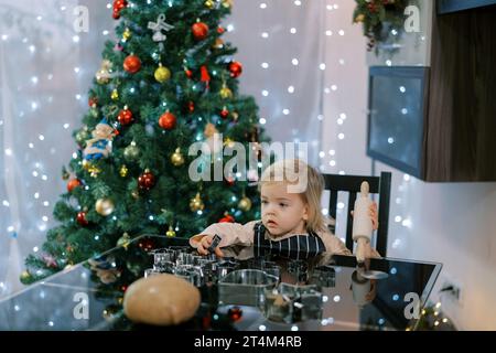 La bambina siede a un tavolo con un mattarello in mano vicino alle taglierine per biscotti e un impasto Foto Stock