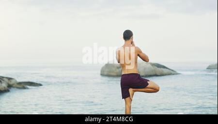 Calma la mente e l'anima parlerà. Foto retrovisiva di un uomo irriconoscibile in piedi e facendo yoga da solo vicino all'oceano durante una giornata di nuvoloso. Foto Stock