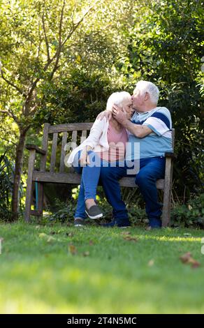 Felice coppia di anziani caucasici seduti sulla panchina, abbracciati e baciati nel giardino soleggiato, spazio fotocopie Foto Stock