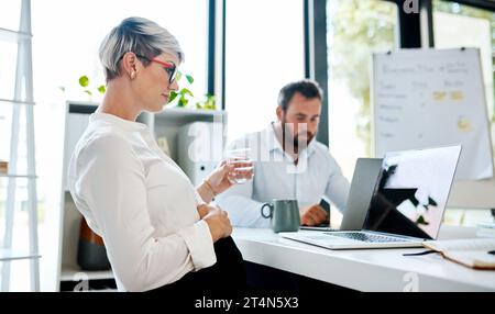 Si prende cura degli affari prima che vada in congedo. una donna d'affari incinta che beve acqua mentre lavora su un computer portatile in un ufficio con il suo collega in Foto Stock