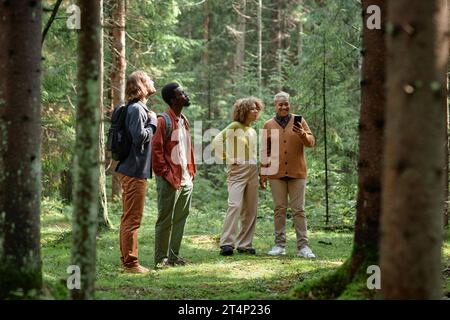 Un gruppo di giovani amici che usano lo smartphone durante le escursioni nella foresta, si trovano sul prato e si godono la natura Foto Stock