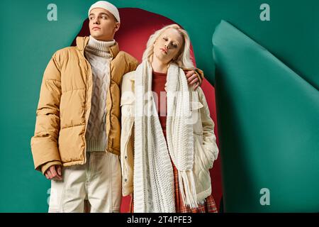 giovane uomo con berretto che abbraccia la donna e in piedi nei capospalla invernali di colore rosso con sfondo turchese Foto Stock