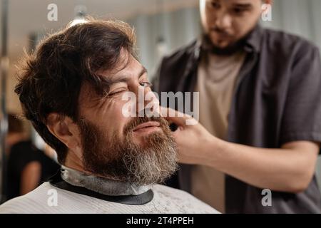 Uomo barbuto che taglia i capelli in barbiere mentre lo stilista lavora con i capelli Foto Stock
